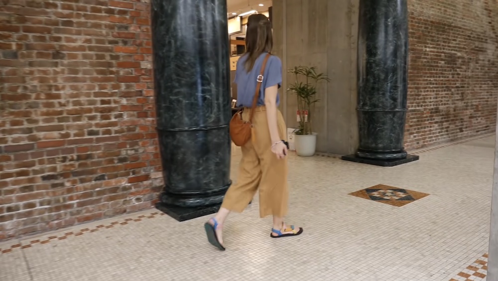 Audrey Bergner Walking Through Historic Brick Interior With Black Columns at Kanemori Warehouses, Hakodate, Japan Audrey Bergner walking through a corridor inside the Kanemori Red Brick Warehouses in Hakodate, Hokkaido, Japan, past exposed red-brick walls, large black columns and a tiled floor with decorative patterns.