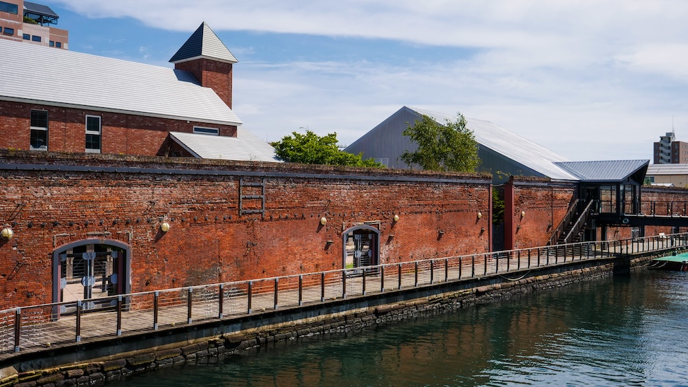 Kanemori Red Brick Warehouses Canal-Side Architecture and Waterfront Walkway in Hakodate, Hokkaido, Japan Historic red-brick warehouse buildings along a canal at Kanemori in Hakodate, Hokkaido, Japan, showing a long brick facade, waterside wooden walkway, metal railings, white rooftops and reflections in the water.