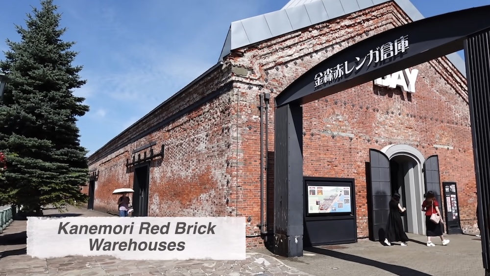 Kanemori Red Brick Warehouses in Hakodate Hokkaido Japan With Historic Brick Architecture and Shopping Entrance Exterior of the Kanemori Red Brick Warehouses in Hakodate, Hokkaido, Japan, showing weathered red brick walls, an arched shopping entrance with Japanese signage, pedestrians and a tall evergreen tree under blue skies.