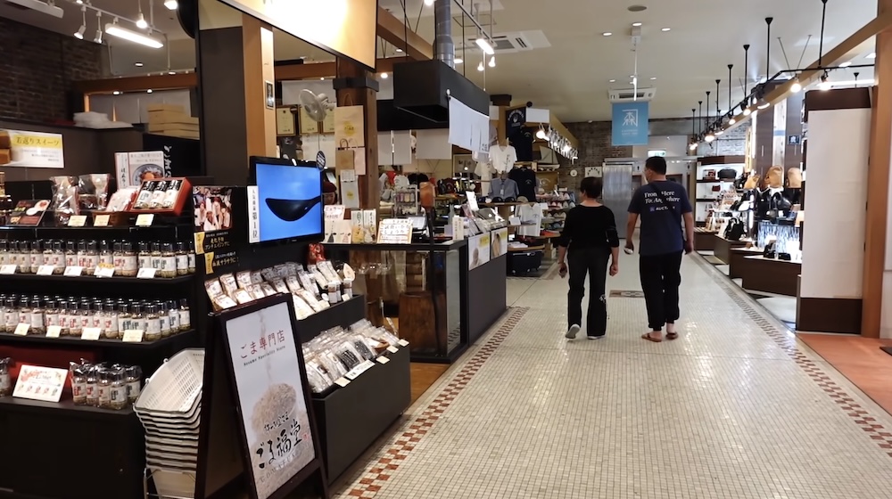 Interior shopping arcade at the Kanemori Red Brick Warehouses in Hakodate, Hokkaido, Japan, with souvenir displays, packaged Japanese foods, wooden shopfronts, overhead lighting and visitors walking along the tiled corridor.