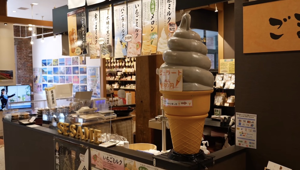 Sesame Soft-Serve Ice Cream Shop and Japanese Dessert Display at Kanemori Red Brick Warehouses, Hakodate, Japan Ice cream counter inside the Kanemori Red Brick Warehouses in Hakodate, Hokkaido, Japan, featuring a large grey soft-serve cone display, sesame signage, Japanese flavour menus and packaged sweets behind glass.