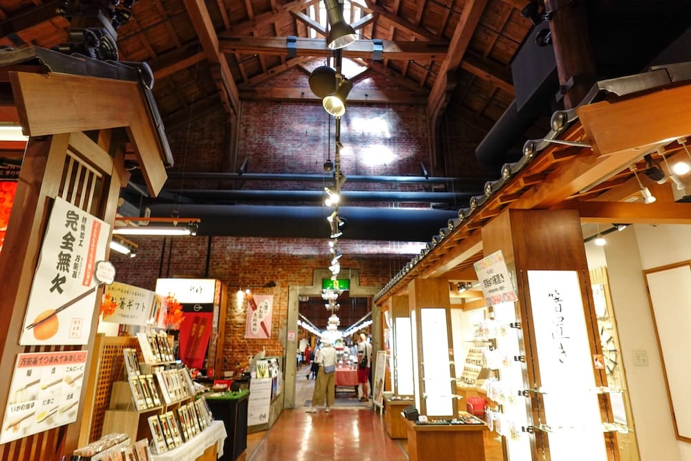 Interior of the Kanemori Red Brick Warehouses in Hakodate, Hokkaido, Japan, showing exposed brick walls, timber roof beams, illuminated shop displays and visitors walking through the commercial arcade.