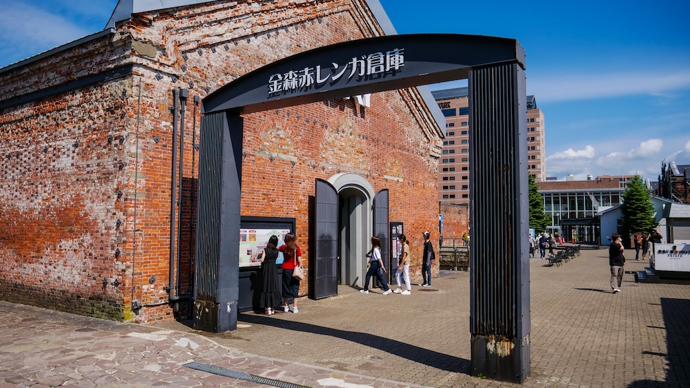 Kanemori Red Brick Warehouses Entrance Arch and Historic Waterfront Buildings in Hakodate, Hokkaido, Japan Entrance to the Kanemori Red Brick Warehouses in Hakodate, Hokkaido, Japan, featuring a black archway with Japanese lettering, weathered red brick buildings and visitors walking through the sunny waterfront complex.