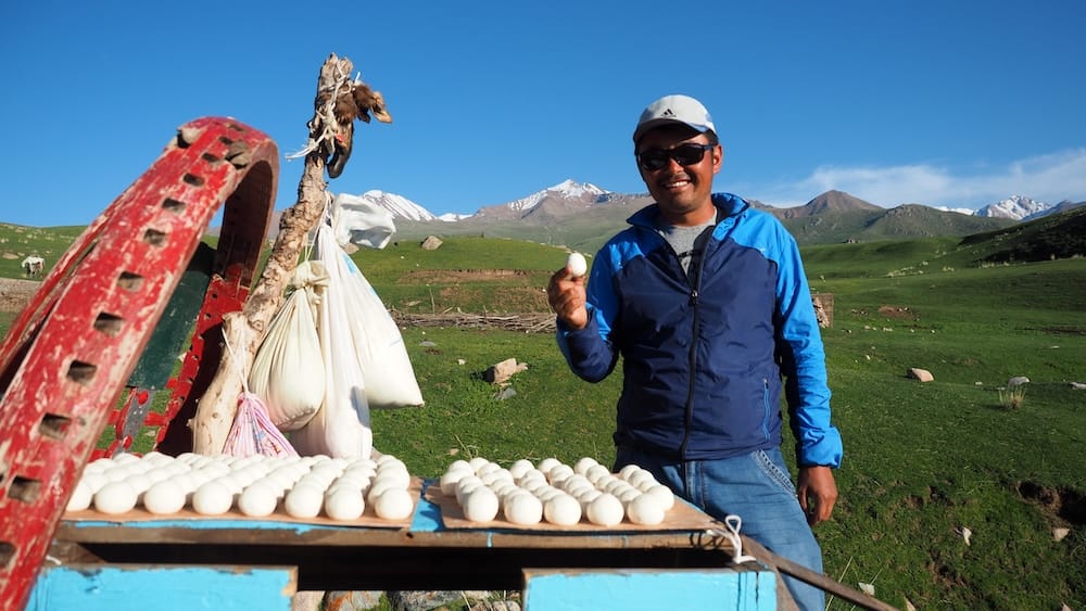 Our Kyrgyz horse-trekking guide holds a ball of traditional kurut beside rows of dried dairy snacks displayed on a wooden cart, with green mountain pastures and snow-capped peaks behind him in Kyrgyzstan.
