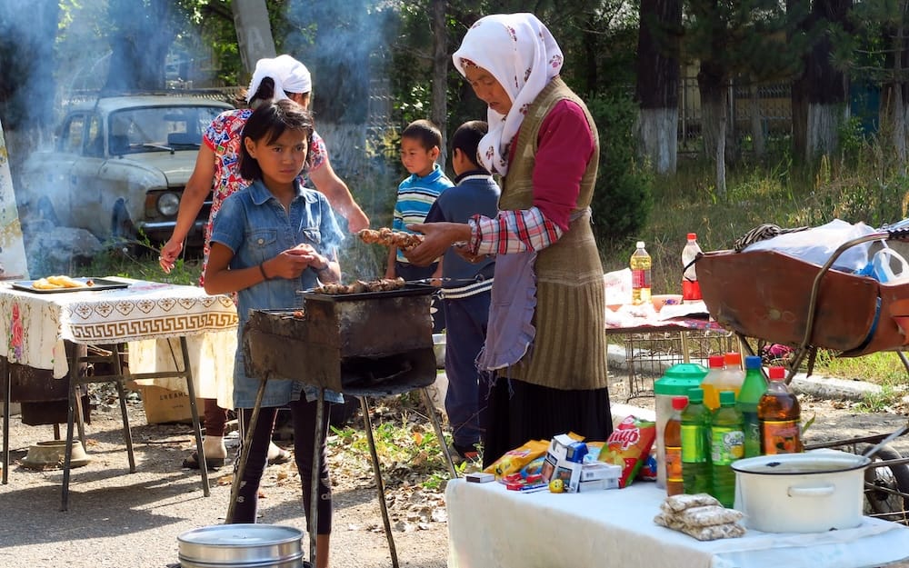 A woman in a headscarf grills Kyrgyz shashlik skewers over a smoky charcoal brazier at an outdoor food stall in Kyrgyzstan, while a young girl stands beside the grill and tables hold drinks and cooking supplies.
