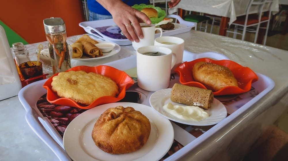 A tray of Kyrgyz yurt snacks and tea includes fried pastries, rolled pancakes, a baked bun, sour cream and several cups, showing the generous assortment of breads and dairy commonly served to guests in Kyrgyzstan.
