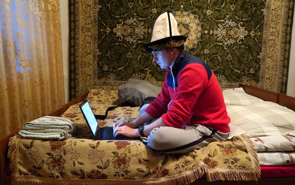 Kyrgyzstan travel scene inside a guesthouse, with Nomadic Samuel sitting on a bed using a laptop while wearing a traditional Kyrgyz felt hat, surrounded by patterned textiles during an early digital-nomad moment.