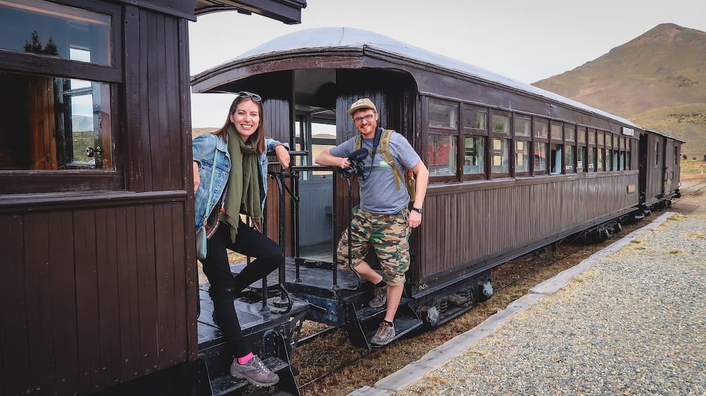 La Trochita steam train carriage, Esquel Station, Chubut, Argentine Patagonia — Samuel Jeffery and Audrey Bergner Audrey Bergner and Samuel Jeffery smiling from the carriage steps of La Trochita, the historic Old Patagonian Express steam train, at Esquel station in Chubut, Argentine Patagonia, with steppe hills behind them.