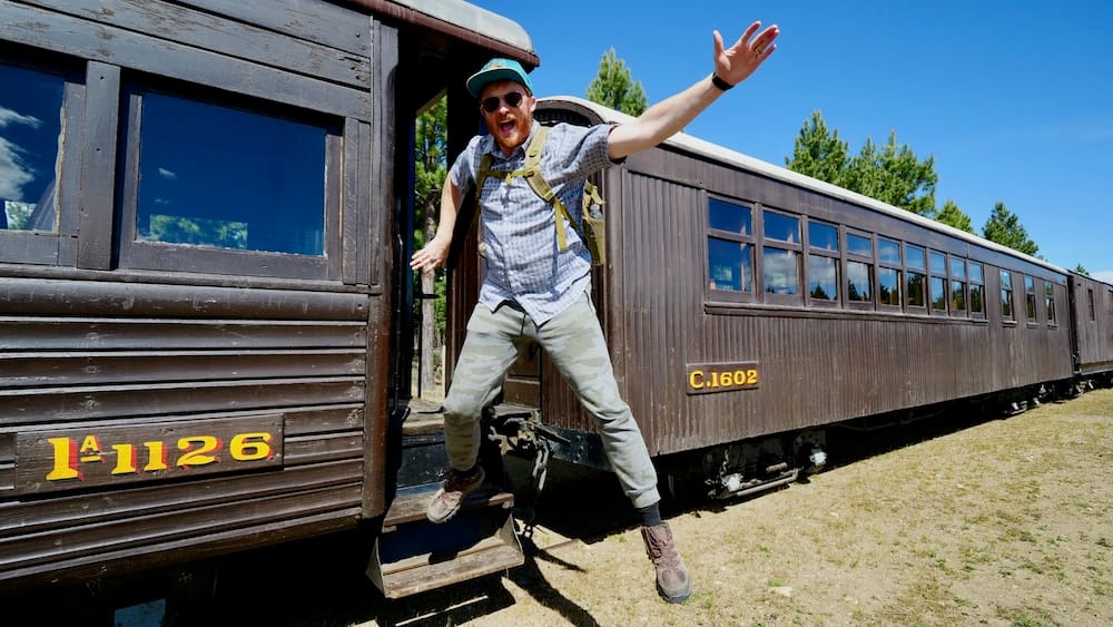 Samuel Jeffery leaps from the steps of a wooden La Trochita heritage carriage in El Maitén, Chubut, Patagonia, Argentina, beneath a clear blue sky beside the historic narrow-gauge train.