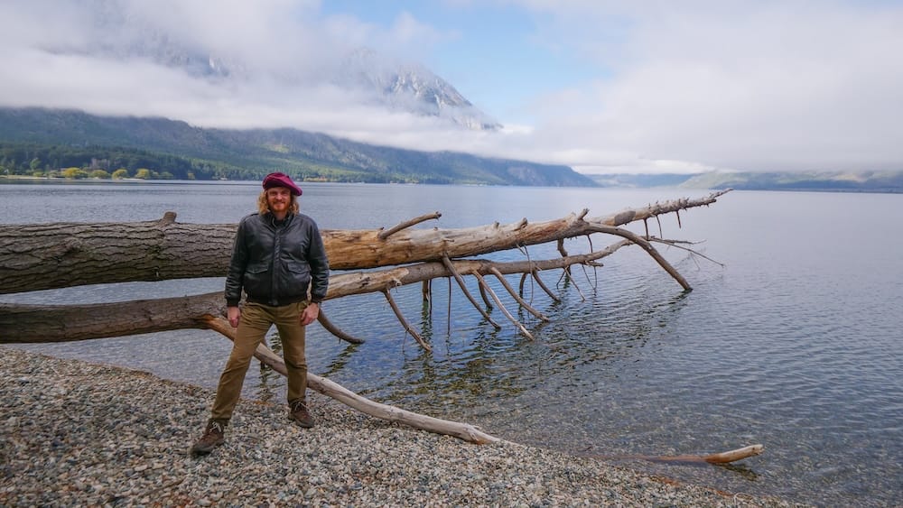 Lago Epuyen Patagonia Argentina with Samuel Jeffery by mountain lake and driftwood shoreline Samuel Jeffery standing beside driftwood along the rocky shore of Lago Epuyen in Patagonia Argentina with calm water, forested hills, and cloud-covered Andes mountains in the background