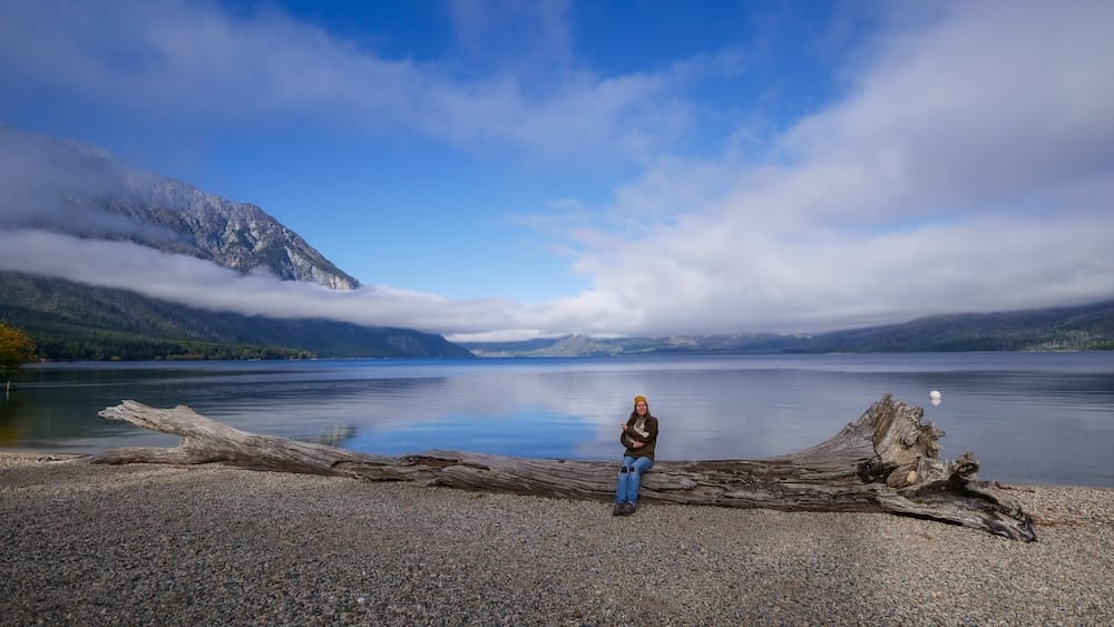 Lago Epuyén Patagonia with Audrey Bergner holding a cat by lakeshore peaceful Andean mountain scenery Lago Epuyén, Chubut Patagonia — Audrey Bergner sitting on a large driftwood log by the lakeshore holding a cat, with calm water and misty Andean mountains behind, capturing the peaceful, intimate moments found in Patagonia’s lesser-visited lakeside towns.
