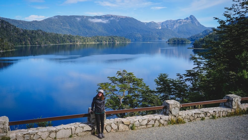 Audrey Bergner at Lago Espejo viewpoint, Seven Lakes Route, Neuquén Province, Patagonia Audrey Bergner stands at a stone-walled viewpoint over Lago Espejo on the Seven Lakes Route in Neuquén Province, Argentine Patagonia, with forested mountains reflected in the still water.