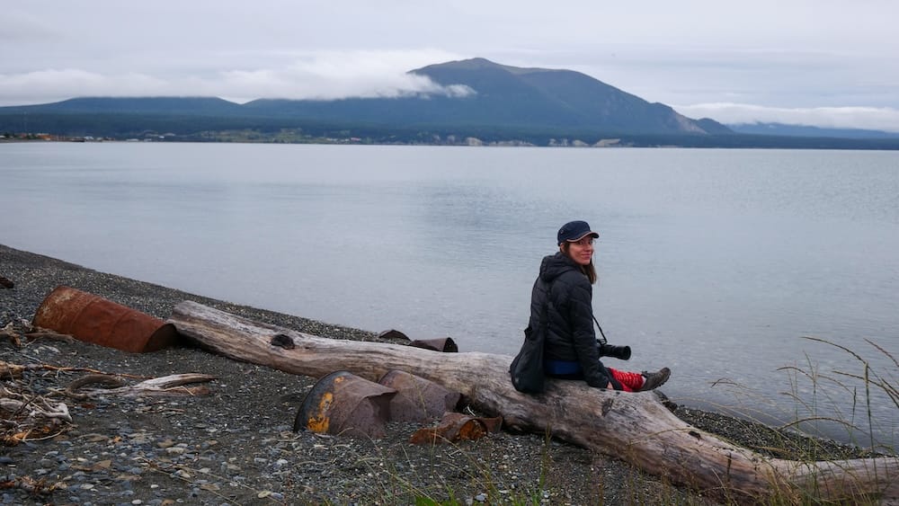 Audrey Bergner sits on driftwood beside the pebbled shore of Lago Fagnano in Tolhuin, Tierra del Fuego, Patagonia, Argentina, with calm water, forested mountains and low cloud behind her.