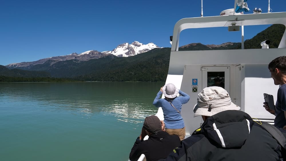 Passengers photograph snowy Andean peaks from a Cruce Andino catamaran crossing Lago Frías near Bariloche, Río Negro, Patagonia, Argentina, over calm turquoise mountain water.