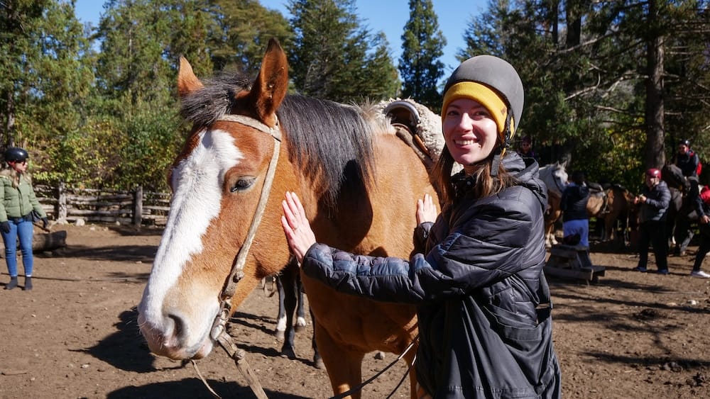 Audrey Bergner stands beside a saddled horse at Lago Gutiérrez in Bariloche, Río Negro, Patagonia, Argentina, wearing a riding helmet in a sunny forest staging area with other riders and horses behind her.