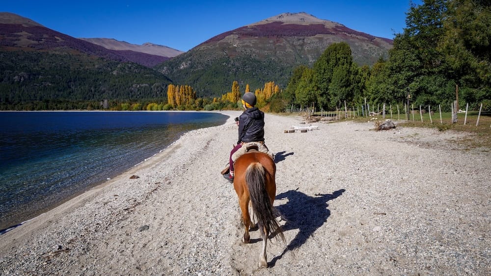 Audrey Bergner cabalga por la orilla de piedras de Lago Gutiérrez cerca de Bariloche, Río Negro, en la Patagonia argentina, con montañas y árboles de colores otoñales enmarcando el lago.