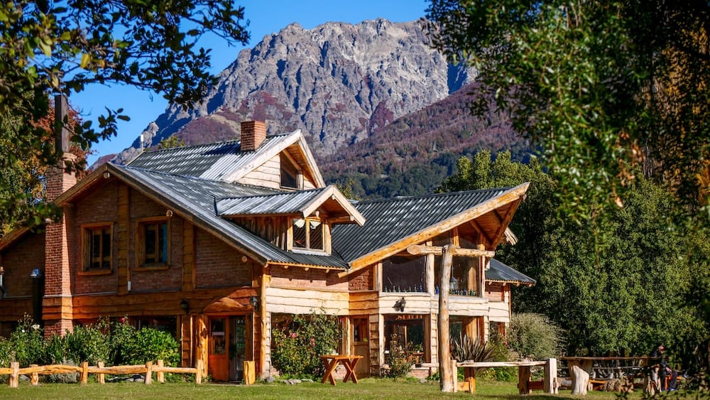 Rustic timber-and-brick chalet at Lago Gutiérrez near Bariloche, Río Negro, framed by forest and sharp mountain peaks beneath a clear blue sky in Argentine Patagonia.