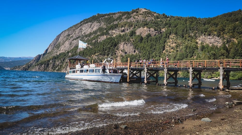 A tour catamaran boards passengers at a wooden pier on Lago Lacar near San Martín de los Andes, Neuquén, Argentine Patagonia, with forested mountains rising behind the water.