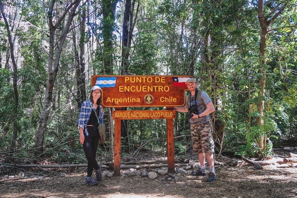 Argentina Chile border at Lago Puelo Patagonia with Samuel Jeffery and Audrey Bergner Samuel Jeffery and Audrey Bergner standing beside Argentina Chile border sign at Lago Puelo National Park in Patagonia surrounded by forest, highlighting the region’s two-country geography and crossing points