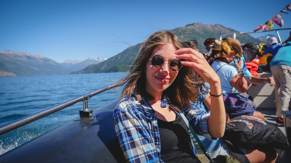 Audrey Bergner sits aboard a boat on Lago Puelo in Chubut, Patagonia, Argentina, with deep blue glacial water, forested mountain slopes and fellow passengers visible under a clear sky.