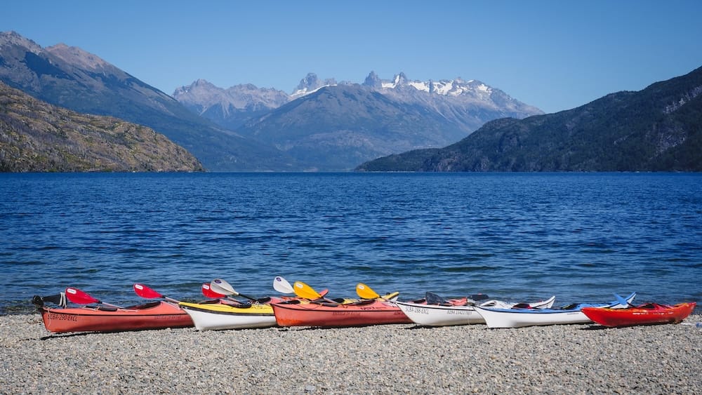 Colourful kayaks rest on the pebble shore of Lago Puelo in Chubut, Patagonia, Argentina, beside deep blue water with forested hills and jagged Andean peaks beneath a clear sky.