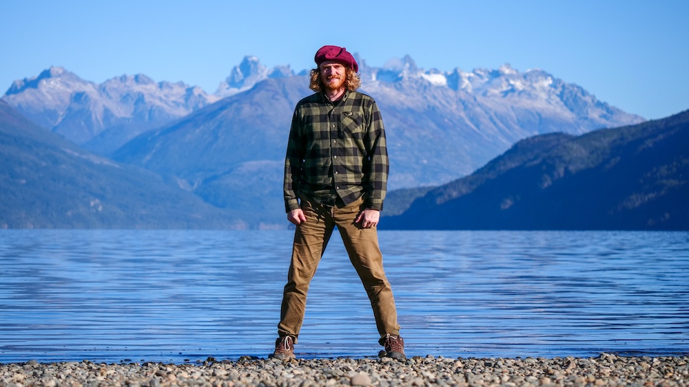 Lago Puelo, Chubut, Patagonia, Argentina mountain-lake scene, with Nomadic Samuel standing on a rocky shoreline wearing a beret, framed by bright blue water and rugged peaks during a remote adventure.