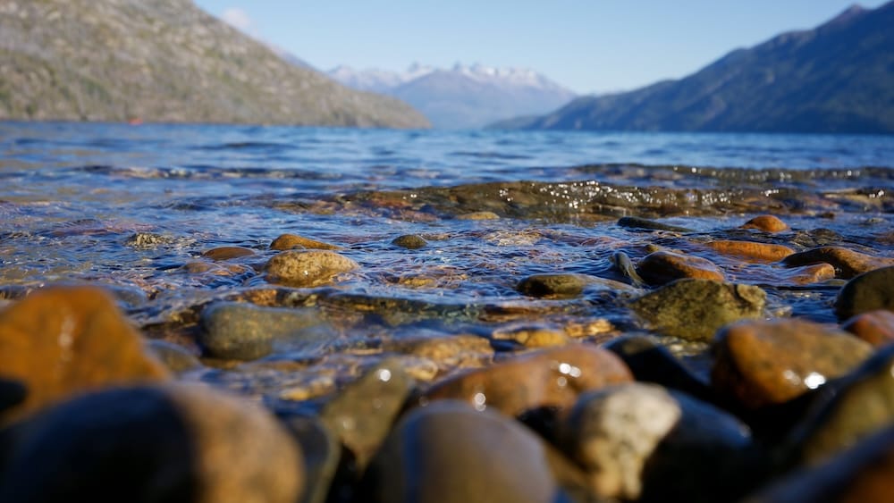 Piedras iluminadas por el sol aparecen bajo el agua clara y poco profunda de la costa del Parque Nacional Lago Puelo, Chubut, Patagonia, Argentina, con el lago azul y montañas desenfocadas al fondo.