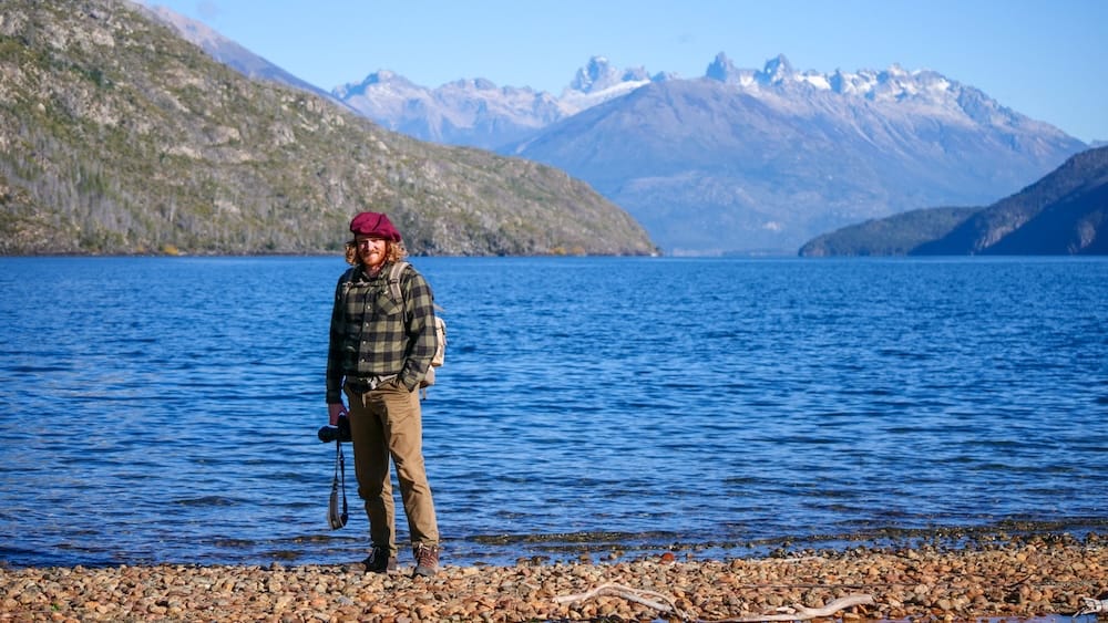 Hiker standing along the rocky shores of Lago Puelo in Patagonia under clear blue skies, where calm-looking conditions can still bring cold air, sudden wind shifts, and microclimate changes typical of autumn and shoulder season travel.
