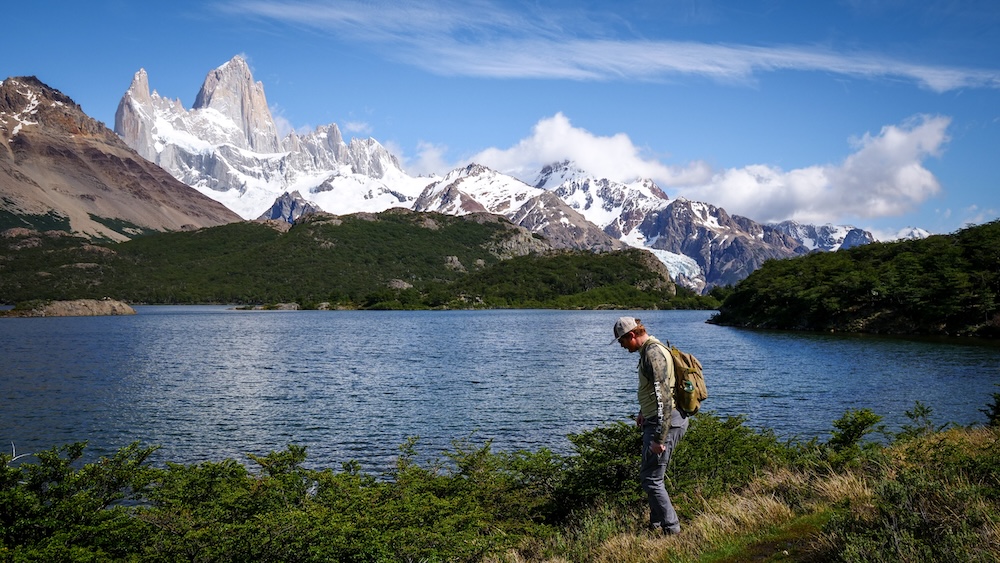 Laguna Capri in El Chaltén, Patagonia, with Mount Fitz Roy towering behind, as Nomadic Samuel hikes along the blue lake shore on a day trip from El Calafate, showing an easy Los Glaciares National Park trail under clear skies.