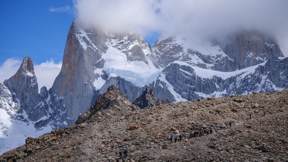 Laguna de los Tres final kilometer in El Chaltén, Patagonia, Argentina, showing tiny hikers on the rocky moraine beneath the towering Fitz Roy peaks as moody clouds roll in, highlighting fast-changing mountain weather.