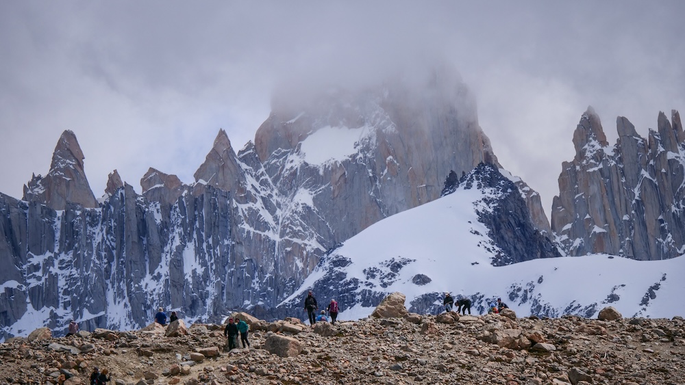 Laguna de los Tres final kilometer in El Chaltén, Patagonia, Argentina, showing tiny hikers crossing the rocky moraine beneath the towering Fitz Roy peaks, highlighting the scale and challenge of the brutal last climb.
