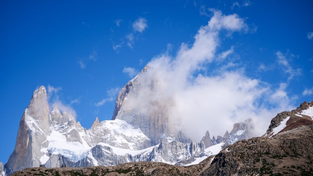 Laguna de los Tres hike, El Chaltén, Patagonia: dramatic Fitz Roy granite spires rise above snowy ridges and rocky terrain under a deep blue sky, with clouds drifting across the peaks during one of the most iconic viewpoints in Los Glaciares National Park.

