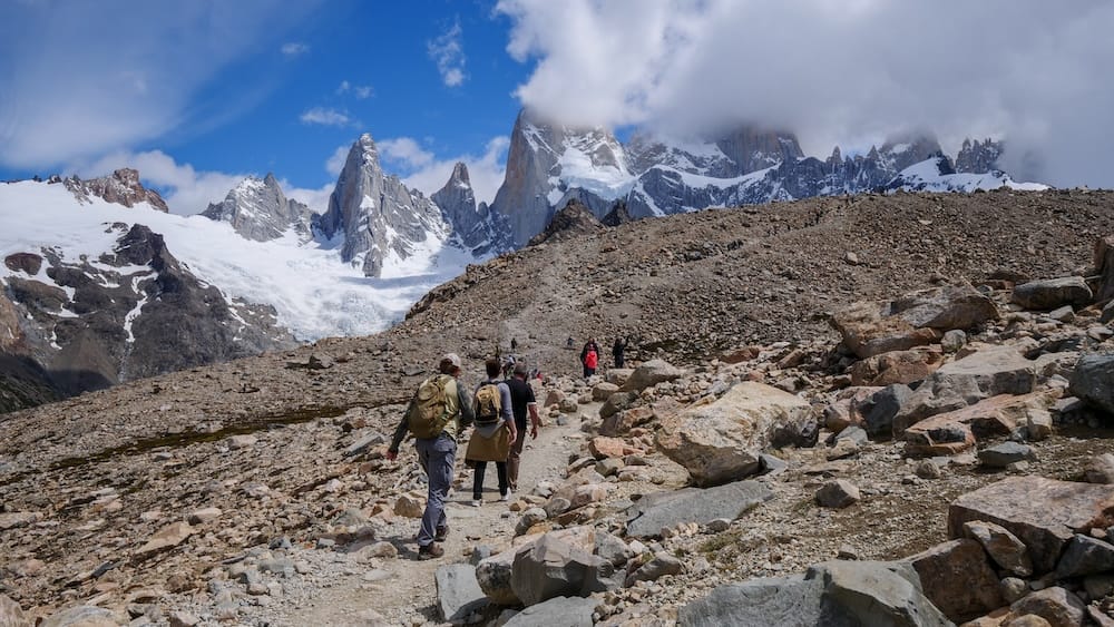 Rocky Laguna de los Tres Approach Near El Chaltén, Patagonia, Argentina, with Samuel Jeffery Samuel Jeffery hikes with other trekkers across a steep, rocky moraine toward the snow-covered Mount Fitz Roy massif near El Chaltén, Santa Cruz, Patagonia, Argentina, beneath shifting cloud and blue sky.