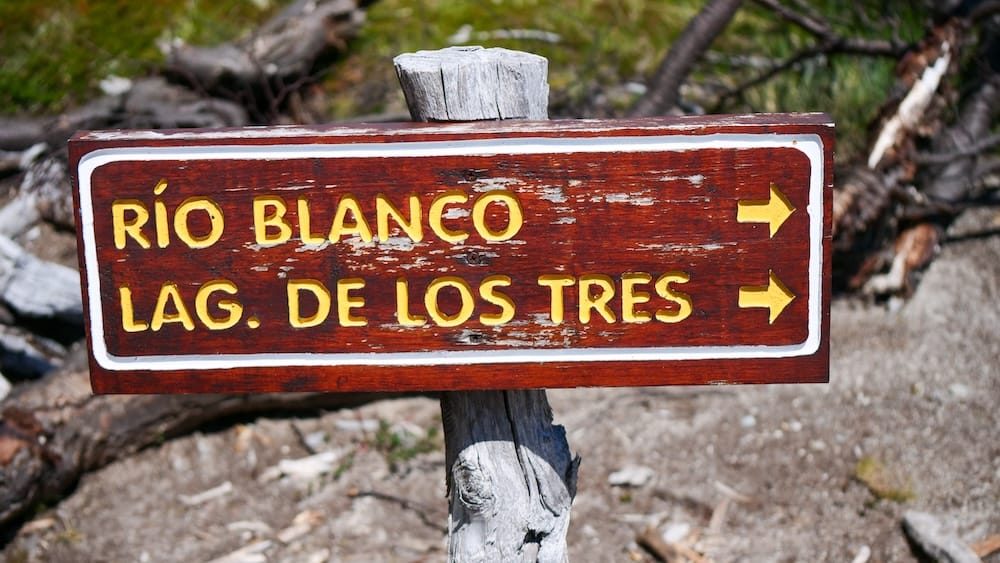 A weathered wooden trail sign points toward Río Blanco and Laguna de los Tres near El Chaltén, Santa Cruz, Patagonia, Argentina, marking the hiking route through rocky forest terrain.