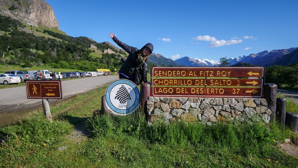 Laguna de los Tres trailhead in El Chaltén, Patagonia, as Audrey Bergner stands full of enthusiasm beside trail signs pointing toward Fitz Roy, ready to begin the iconic Laguna de los Tres hike in Argentina’s trekking capital.
