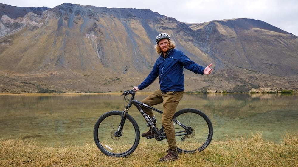 Samuel Jeffery pauses with a mountain bike beside a clear lake near Esquel, Chubut, Patagonia, Argentina, with steep rocky Andean slopes rising across the water beneath an overcast sky.