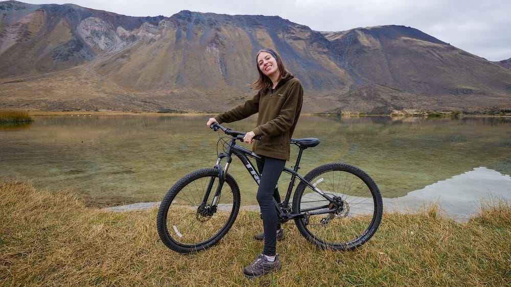 Audrey Bergner at Laguna La Zeta in Esquel Patagonia with calm autumn lake reflections and mountain backdrop Laguna La Zeta near Esquel in Chubut, Argentina during autumn, with Audrey Bergner standing beside a bike on the lakeshore, clear glacial water reflecting rugged Patagonian mountain slopes under calm shoulder season conditions.