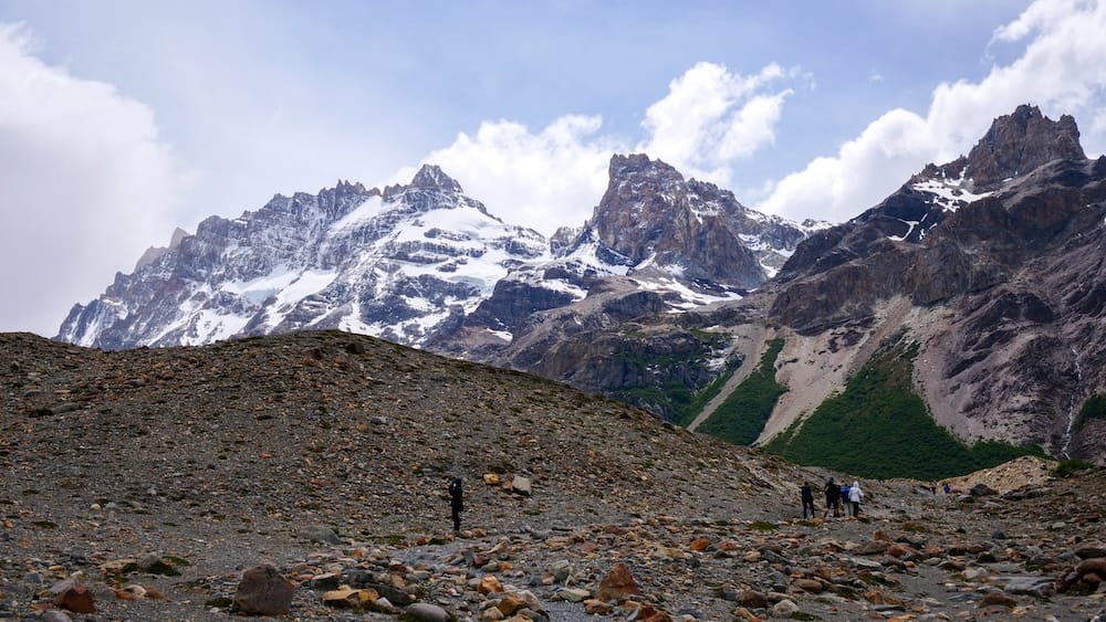 Laguna Torre hike in El Chaltén, Patagonia, Argentina, with hikers crossing a rocky moraine beneath towering, snow-streaked peaks and dramatic mountain walls, highlighting the epic scenery that makes this full-day trek one of the region’s most rewarding routes.
