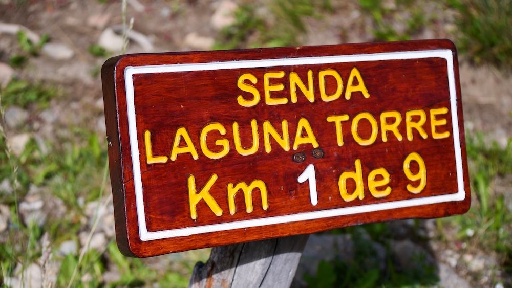 Wooden trail sign reading “Senda Laguna Torre Km 1 de 9” marking the start of the Laguna Torre hike in El Chaltén, Patagonia, Argentina, guiding hikers into the haunted Lenga forest and glacial valley beyond