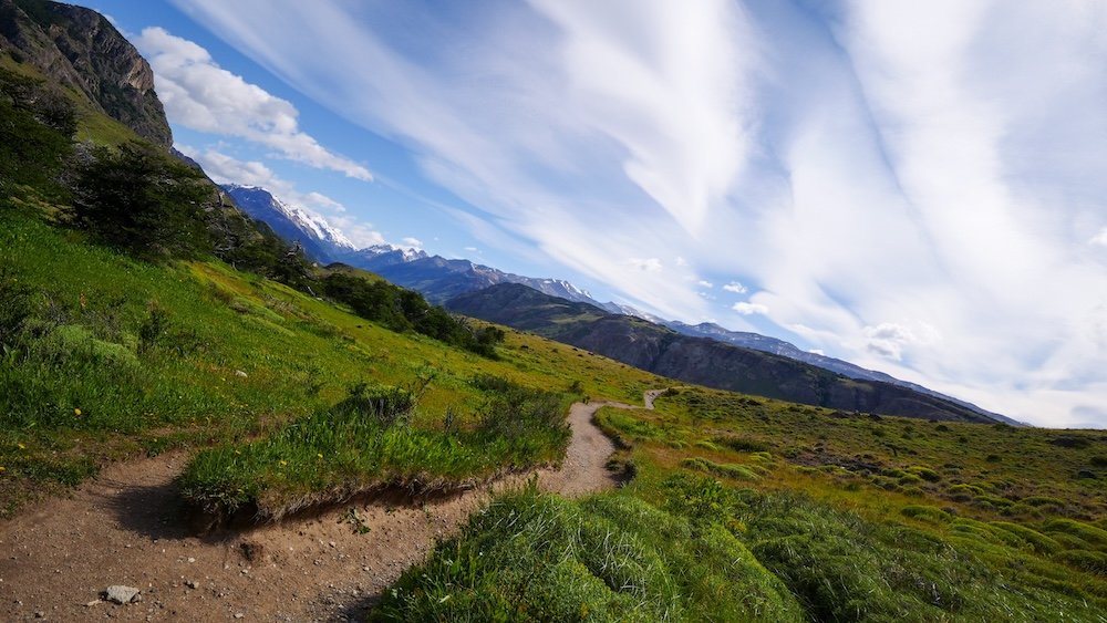 Laguna Torre hike, El Chaltén, Patagonia: a winding dirt trail cuts through green grassland under dramatic streaked clouds, showing the wide-open, windy valley scenery that defines the early stages of this iconic day hike in Los Glaciares National Park.
