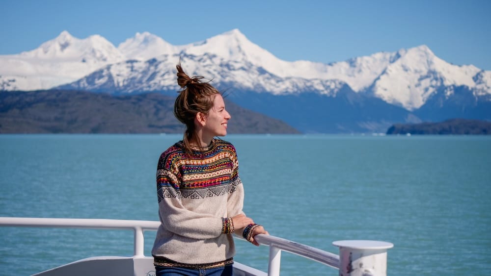 Audrey Bergner stands on a boat deck during a Lake Argentino tour near El Calafate, Santa Cruz, Patagonia, Argentina, with turquoise glacial water and snow-covered mountains rising across the horizon.