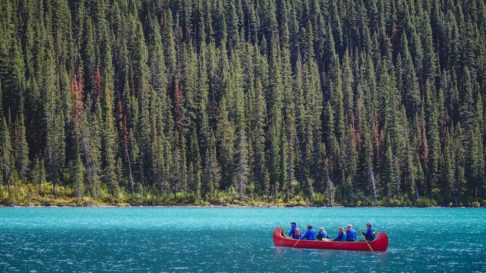 A red canoe carrying a small group paddles across turquoise Lake Louise in Alberta, Canada, with dense evergreen forest rising along the shoreline behind them.