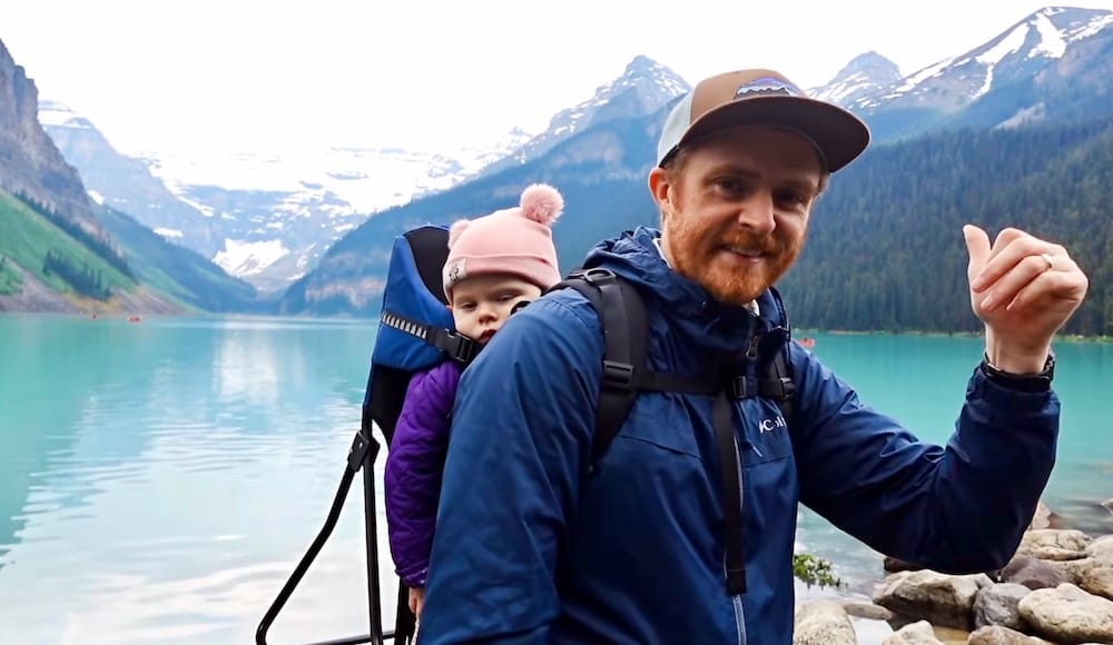 Samuel Jeffery lleva a Aurelia Jeffery en una mochila portabebé junto al agua turquesa de Lake Louise, Alberta, Canadá, con laderas boscosas, montañas nevadas y paisaje glaciar detrás.