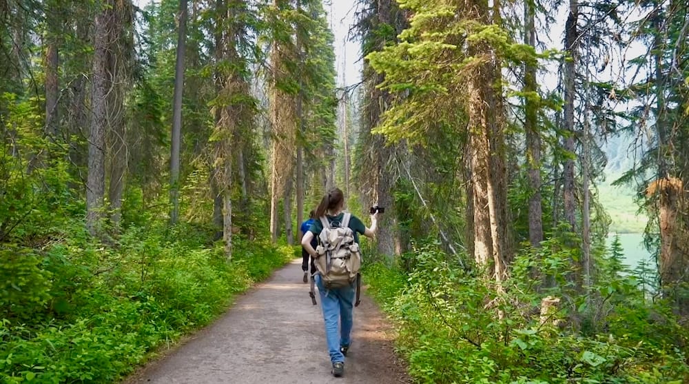 Audrey Bergner hikes along a forest trail at Lake Louise in Alberta, Canada, carrying a backpack and camera with dense evergreen woodland surrounding the path and glimpses of the lake through the trees.