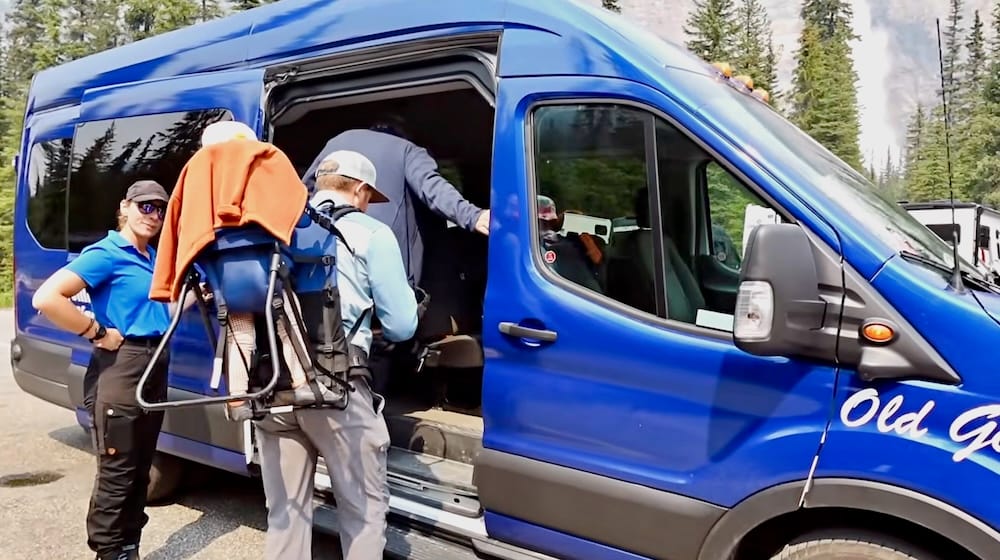 Nuestra familia vuelve a subir a un vehículo azul de excursión privada cerca de Lake Louise, Alberta, Canadá, con una mochila portabebé visible y bosque de coníferas alrededor de la parada.