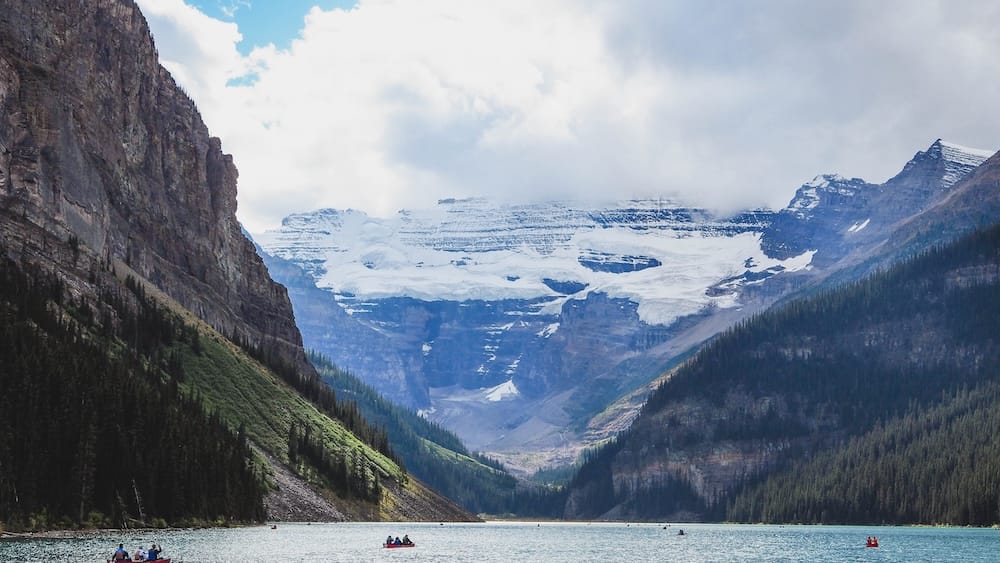 Lake Louise, Alberta, Canadá, con agua turquesa y canoas rojas bajo laderas boscosas empinadas, picos nevados y paisaje glaciar parcialmente cubierto por nubes.