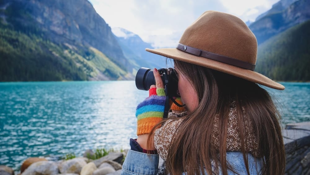 Audrey Bergner photographs turquoise Lake Louise in Alberta, Canada, from the rocky shoreline, with steep forested mountains and pale glacial scenery rising beyond the water.
