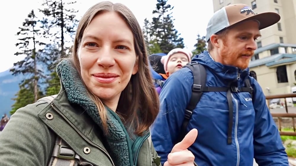 Audrey Bergner sonríe junto a Samuel Jeffery, que levanta el pulgar cerca del Fairmont Chateau en Lake Louise en el Parque Nacional Banff, Alberta, Canadá, con su hija pequeña Aurelia Jeffery dormida en una mochila portabebés entre ellos.
