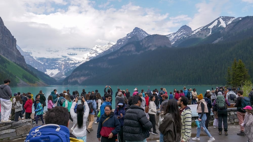 Heavy crowds gathered along the Lake Louise shoreline in Banff National Park, Alberta, Canada, with turquoise water, forested mountain slopes and snowy peaks visible beyond the busy viewing area.