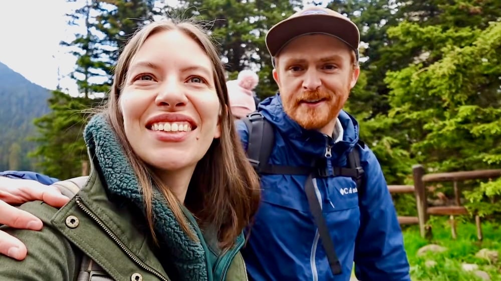 Audrey Bergner and Samuel Jeffery exploring Lake Louise in Banff National Park, Alberta, Canada, with Aurelia Jeffery in a child carrier behind them and evergreen forest surrounding the trail.